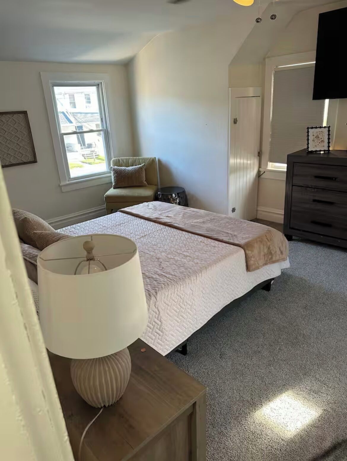 A bedroom with a bed, a bedside table with a lamp, a dresser, a chair, and a window.
