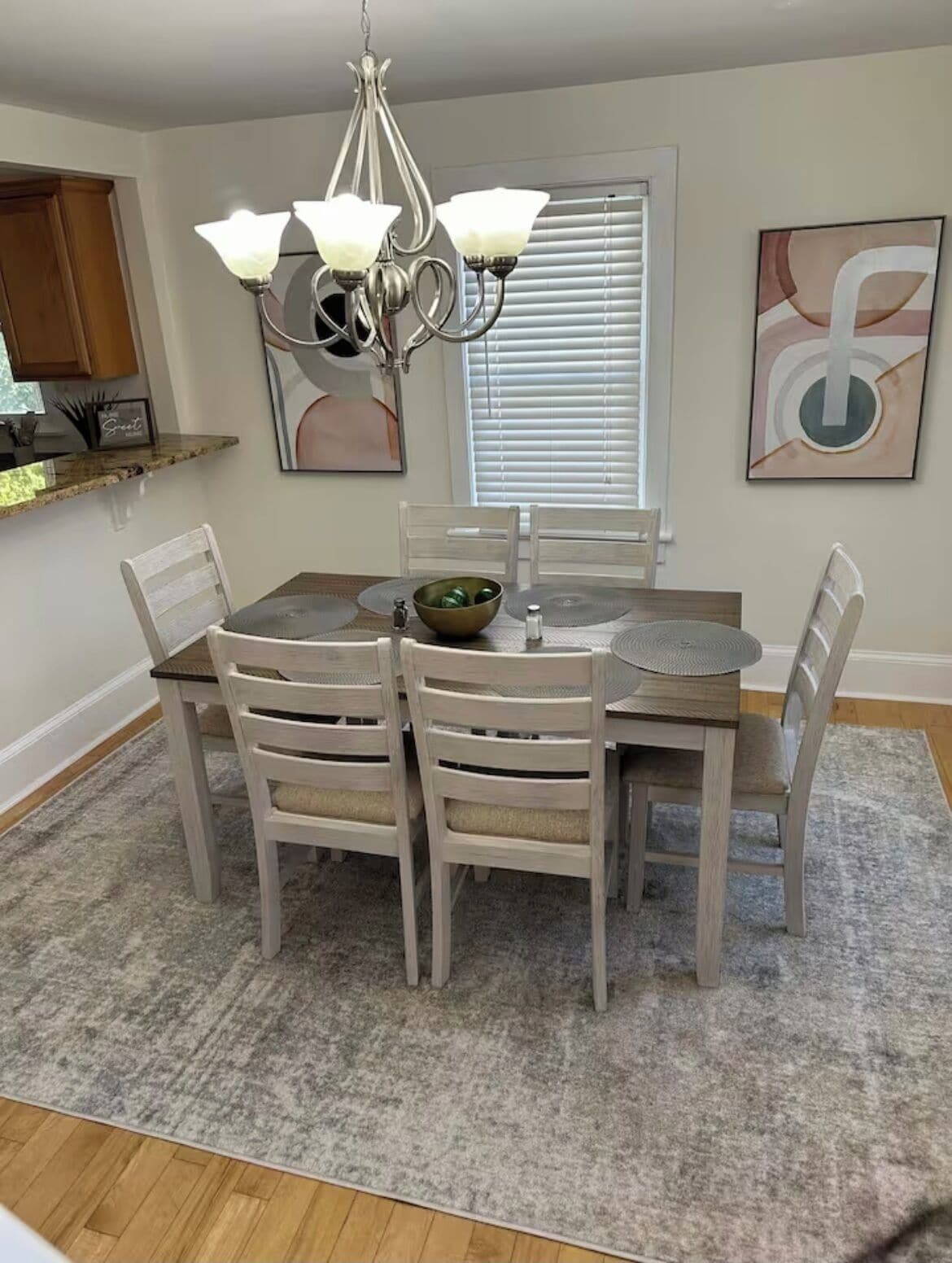 A dining area with a round table, six chairs, a chandelier, a window with blinds, and artwork on the walls.