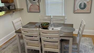 A dining area with a round table, six chairs, a chandelier, a window with blinds, and artwork on the walls.