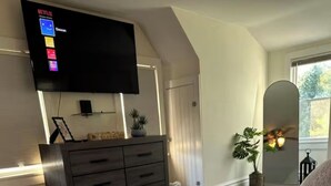 A bedroom with a large bed, a dresser, a flat-screen TV mounted on the wall, and a window with curtains. -  Cozy Cape Cod in lovely Allentown with modern amenities (Allentown)