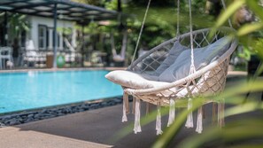 A hanging chair by a pool with a clear view of the pool and a building in the background. - Banana Resort le Petit nid de Caro (Siem Reap)
