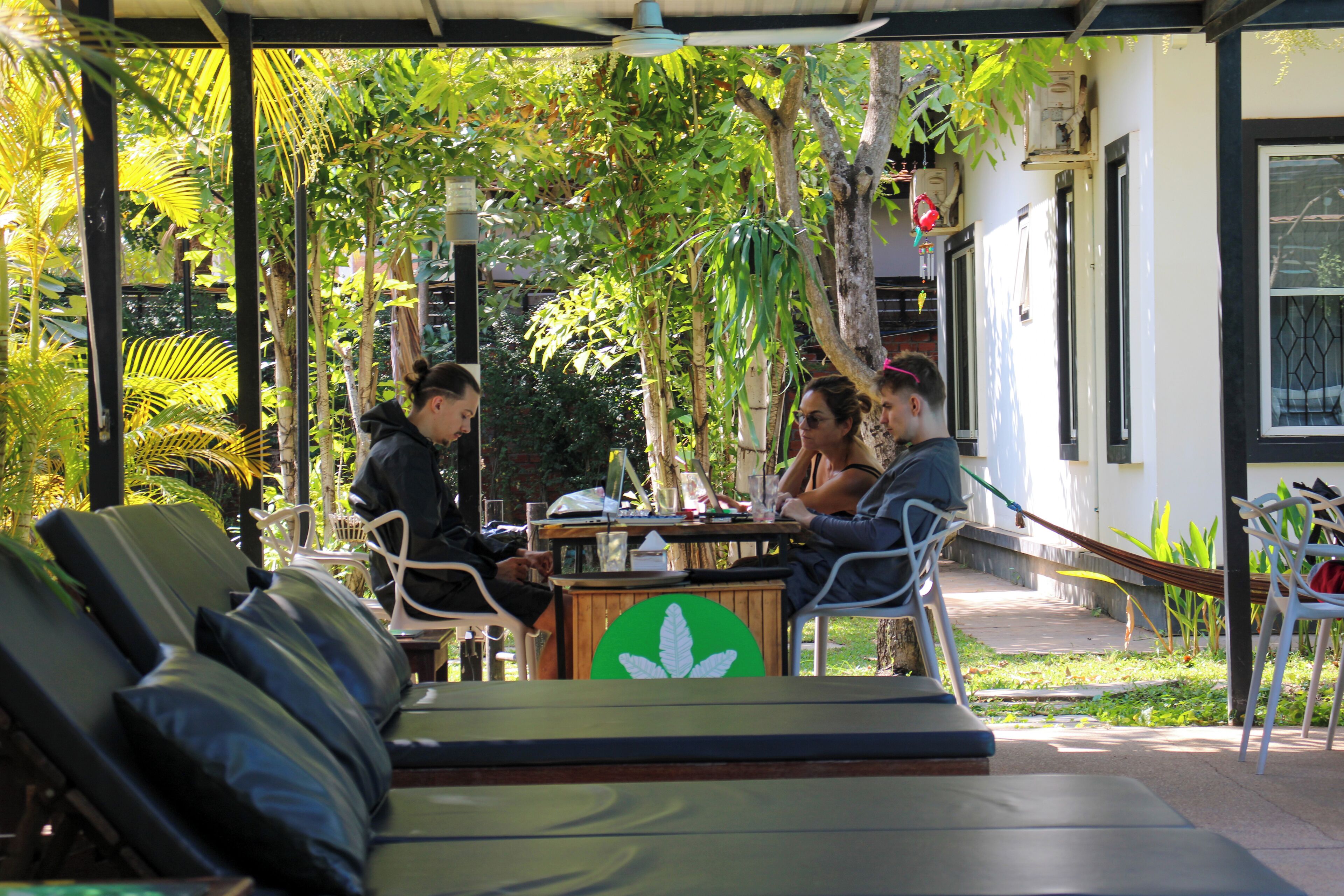 A covered outdoor seating area with lounge chairs and a table, surrounded by greenery and a building with a window.