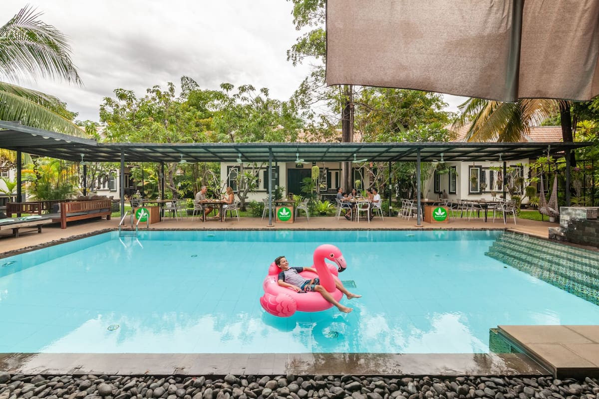 Uma piscina com dois flâmicos cor-de-rosa infláveis, rodeada por áreas de estar cobertas e vegetação.