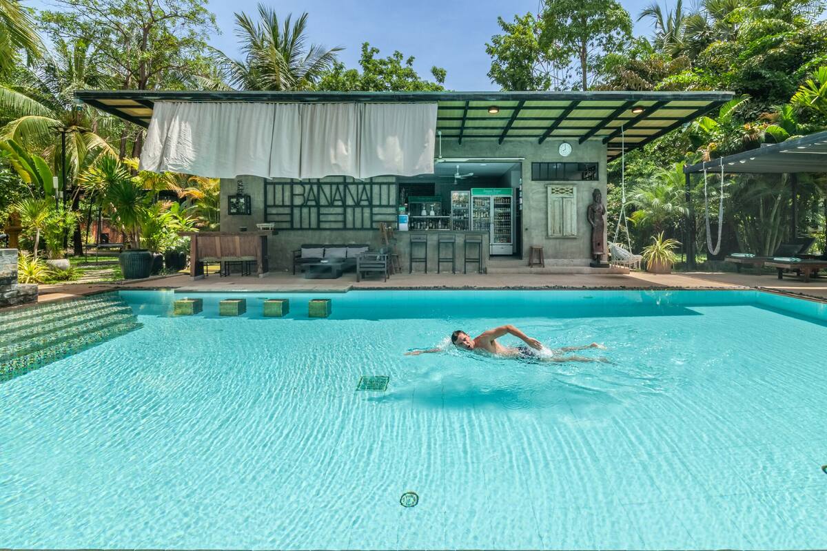 Piscina com uma pessoa nadando, área de bar e espaço coberto para sentar.