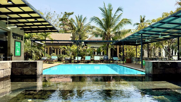 A swimming pool with lounge chairs and umbrellas, surrounded by palm trees and a building with a sign. - Banana Resort le Petit nid de Caro (Siem Reap)