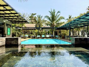 A swimming pool with lounge chairs and umbrellas, surrounded by palm trees and a building with a sign. - Banana Resort le Petit nid de Caro (Siem Reap)