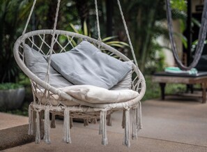A wicker hanging chair with cushions, a wooden bench, and a potted plant in the background. - Banana Resort le Petit nid de Caro (Siem Reap)