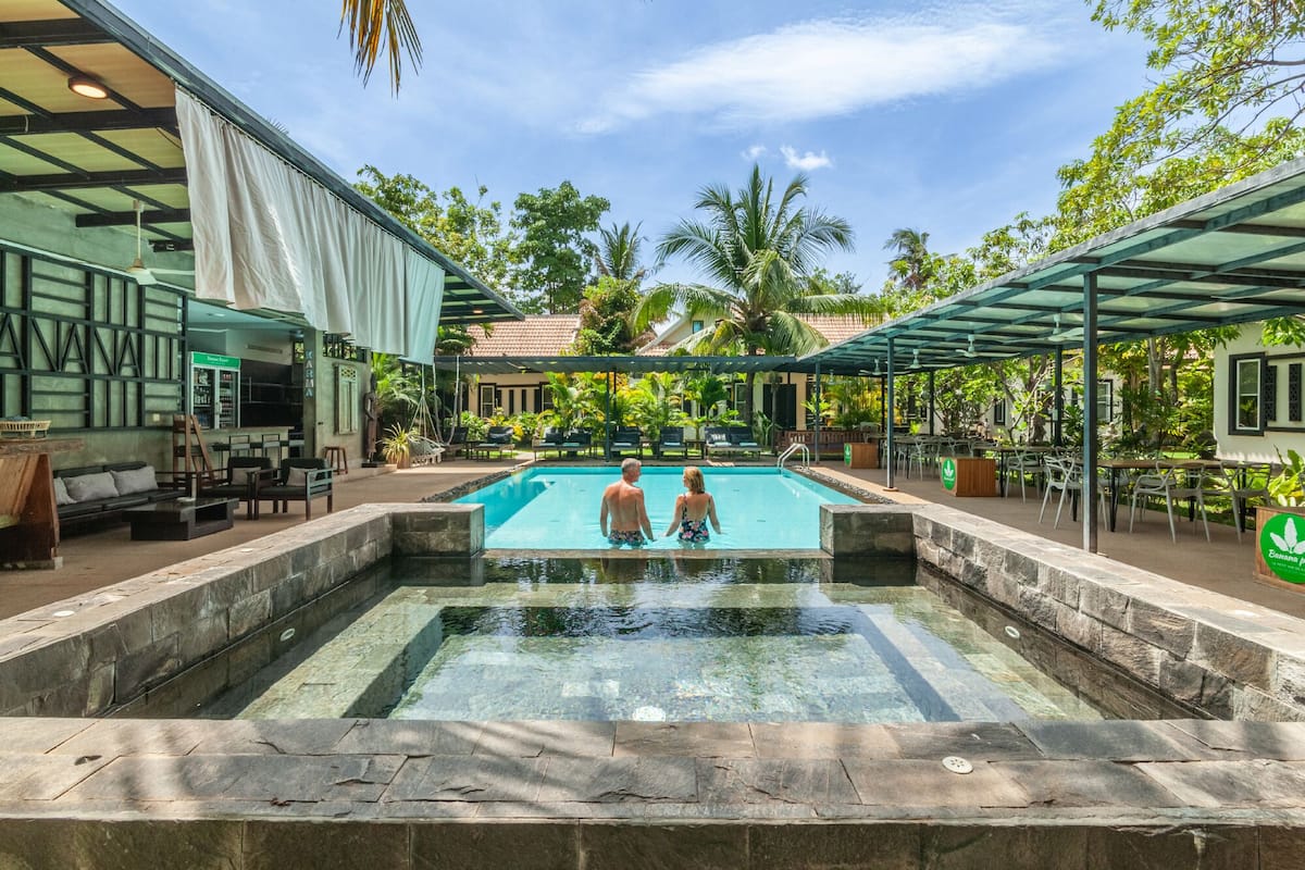 Uma área de piscina com duas pessoas, cadeiras de descanso e um espaço coberto para sentar.