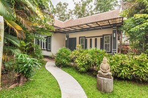 A house with a curved concrete pathway leading to a door, surrounded by lush greenery and a Buddha statue. - Banana Resort le Petit nid de Caro (Siem Reap)