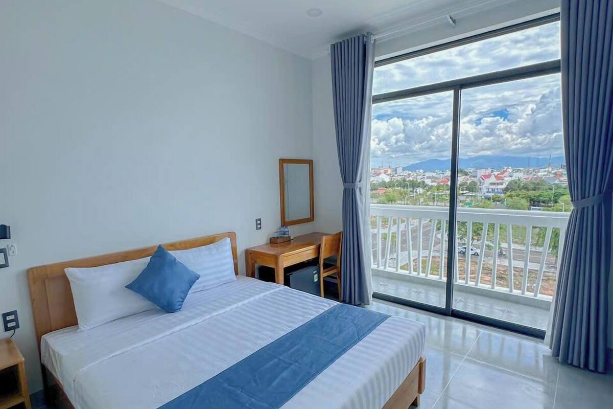 A hotel room with a large bed, a desk, and a view of a cityscape through large windows.
