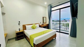 A hotel room with a large bed, a bedside table with a phone, a mirror, and a view of the city through a balcony. - Luna Hotel Phan Thiet (Phu Thuy)
