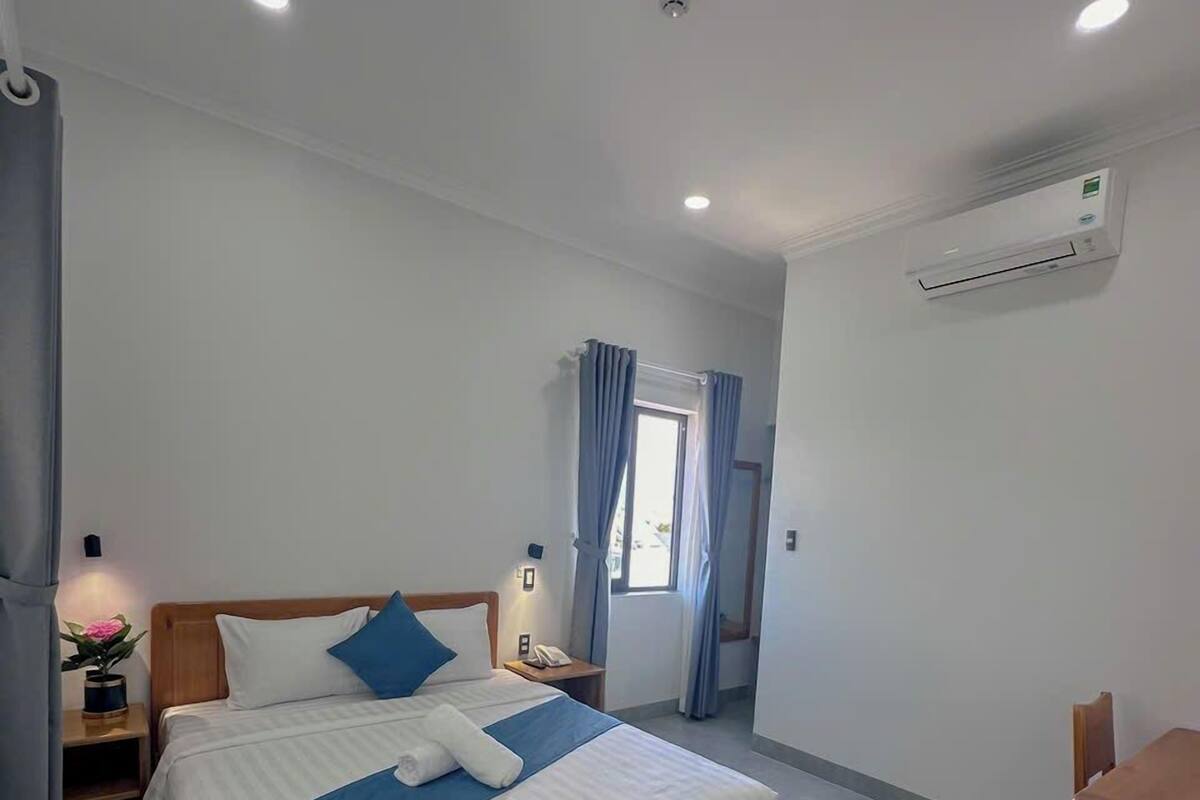 A modern hotel room with a wooden bed, a bedside table with a lamp, a small desk, and a window with curtains.