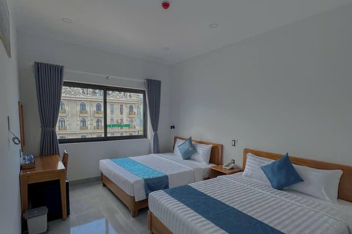 A hotel room with two beds, a desk, a chair, a small trash bin, and a window with a view of a building.