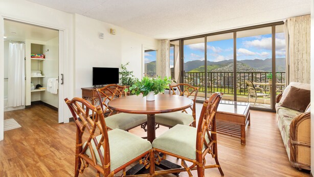 Amplia sala de estar con una mesa redonda de madera rodeada de sillas de mimbre, un televisor de pantalla plana y una planta en maceta. Se aprecia un balcón con vistas panorámicas a la montaña a través de puertas de cristal grandes.