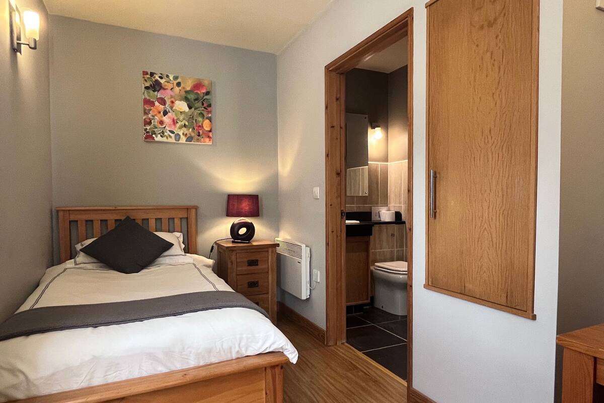 A bedroom with a wooden bed, a nightstand with a lamp, a painting on the wall, and a view into the bathroom.