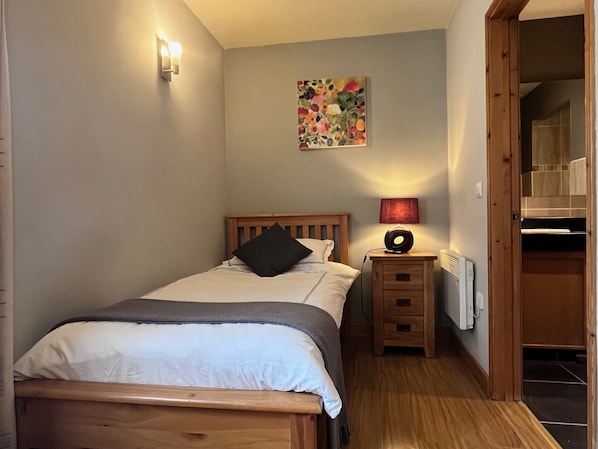 Een slaapkamer met een houten bed, een nachtkastje met een lamp, een radiator, en een deur naar een badkamer.