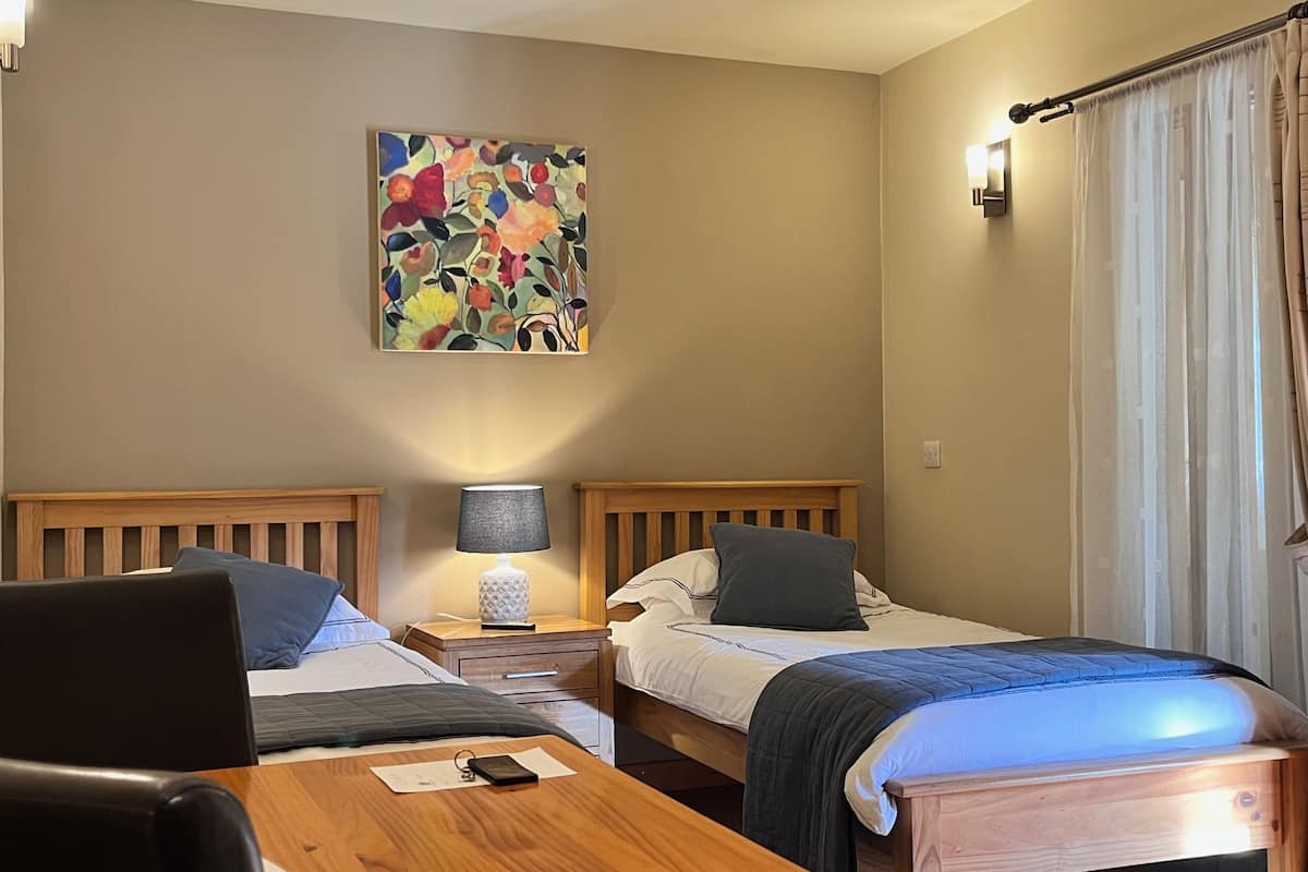 A hotel room with two beds, a desk, a lamp, a painting on the wall, and a window with curtains.