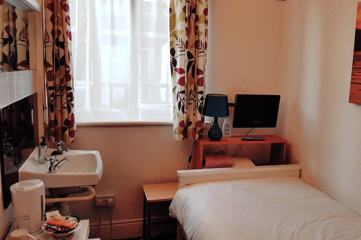 Egy szállodai szoba, amelyben található egy ágy, egy íróasztal számítógéppel, egy mosdókagyló és egy ablak, amelyen függöny van.