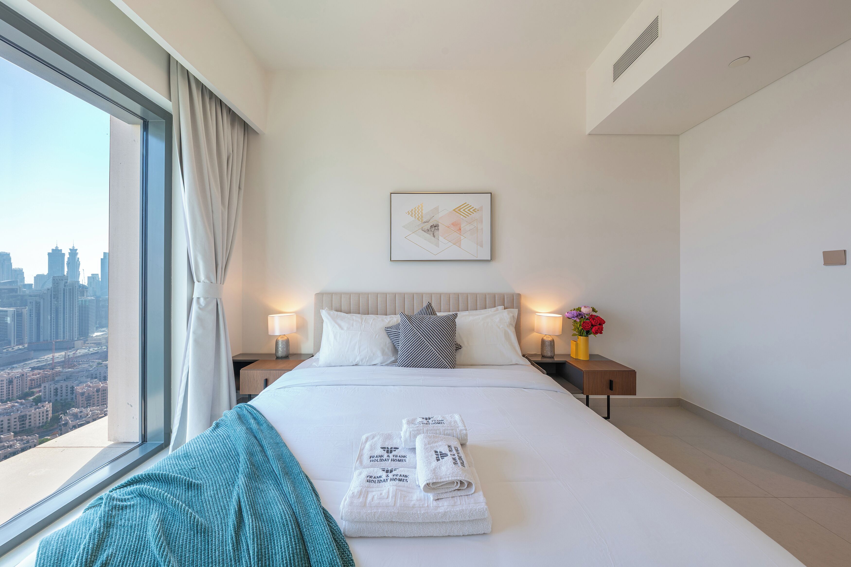 A hotel room with a large bed, a city view, a framed artwork on the wall, and a nightstand with a vase of flowers.