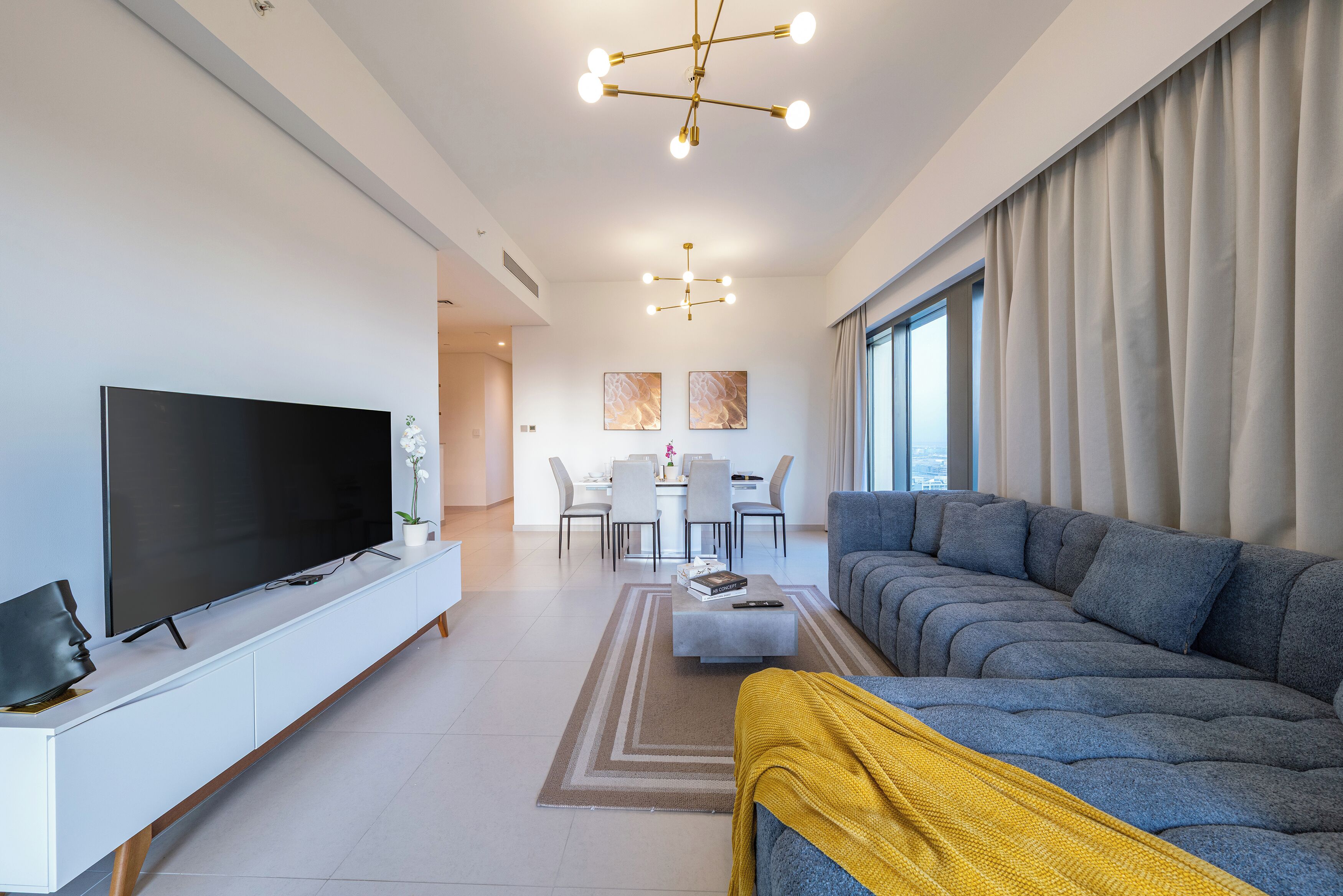 Un salon moderne avec un grand téléviseur à écran plat, un canapé gris, une table basse et un plaid jaune. On aperçoit à l’arrière-plan un coin repas avec une table et des chaises.