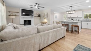 A modern living room with a large sofa, a fireplace, and a kitchen area with white cabinets and a bar stool. - Forest Retreat: Cozy Getaway (Wake Forest)