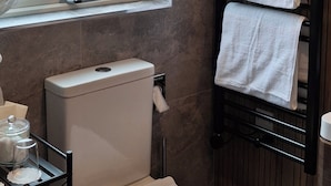 A modern bathroom with a toilet, a towel rack with two towels, a window with a curtain, and a shelf with toiletries. - Quaint studio in Galway, Clarinbridge for a peaceful getaway (Galway, Clarinbridge)
