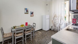 A compact kitchen with a dining table, chairs, a refrigerator, a gas stove, and a window with curtains. - Happy Family Apartment by Wonderful Italy (Genoa)
