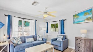 Una sala de estar con un sofá azul, una mesa de centro de madera, un ventilador de techo y un cuadro con una playa.
