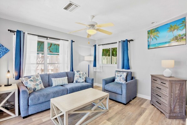 A living room with a blue sofa set, a wooden coffee table, a ceiling fan, and a painting of a beach scene.