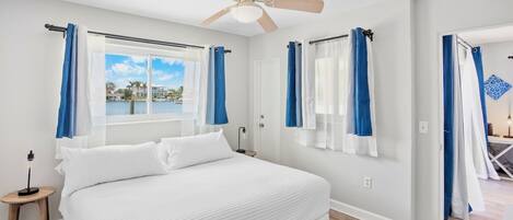 A bedroom with a large bed, a ceiling fan, and a view of a body of water through the window.