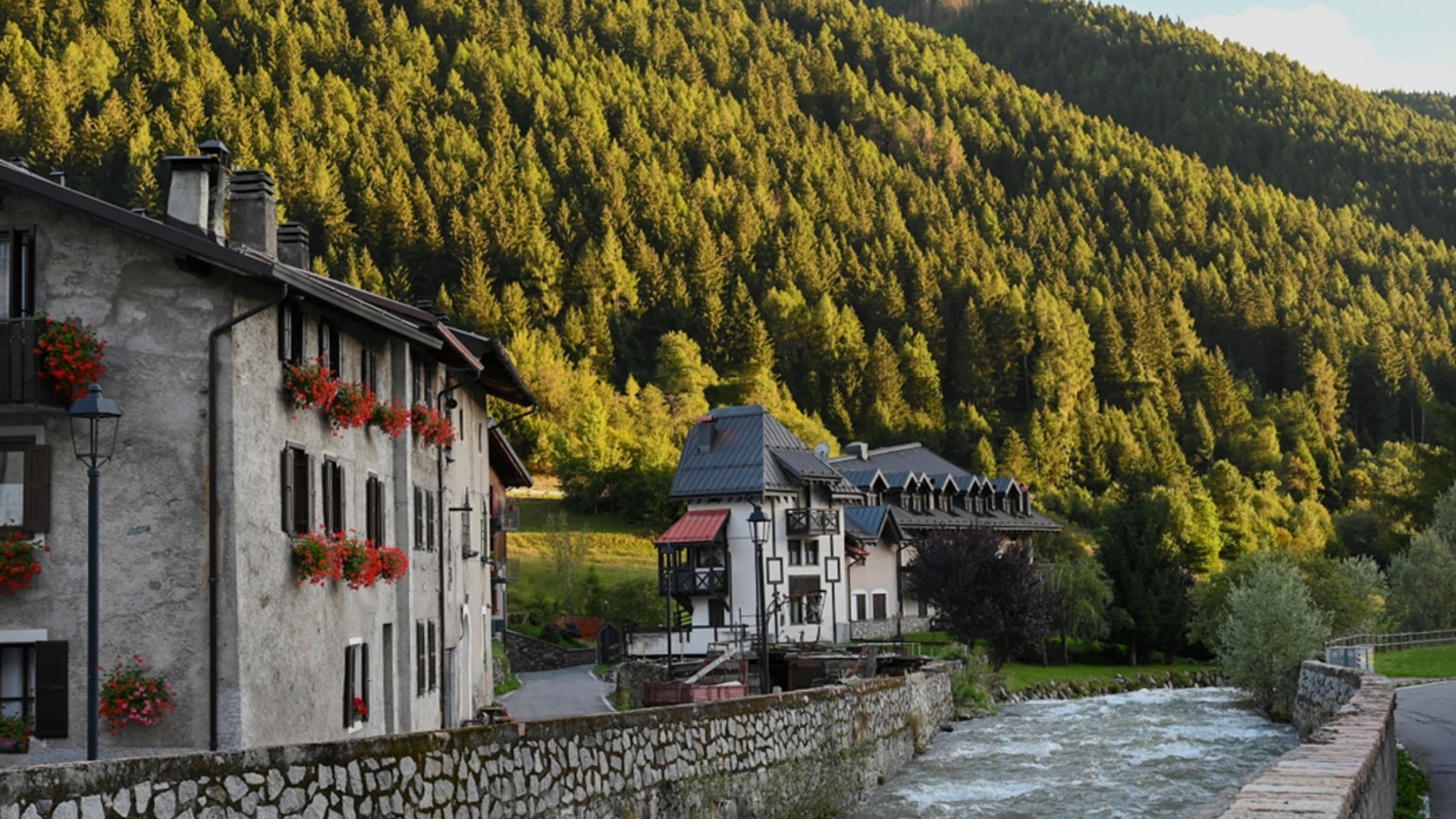 A stone-walled building with a river in the foreground and a forested hill in the background.