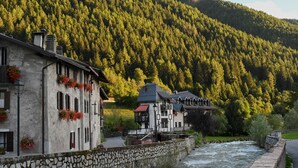 A stone-walled building with a river in the foreground and a forested hill in the background. - Castelpoggio Luxury Apartment (Ponte di legno)