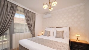 Un dormitorio ordenado con una cama grande, dos mesitas de noche con lámparas, una ventana con cortinas y un equipo de aire acondicionado en la pared.