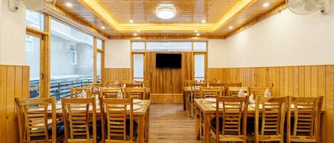 A dining area with wooden tables and chairs, a TV mounted on the wall, and ceiling fans.