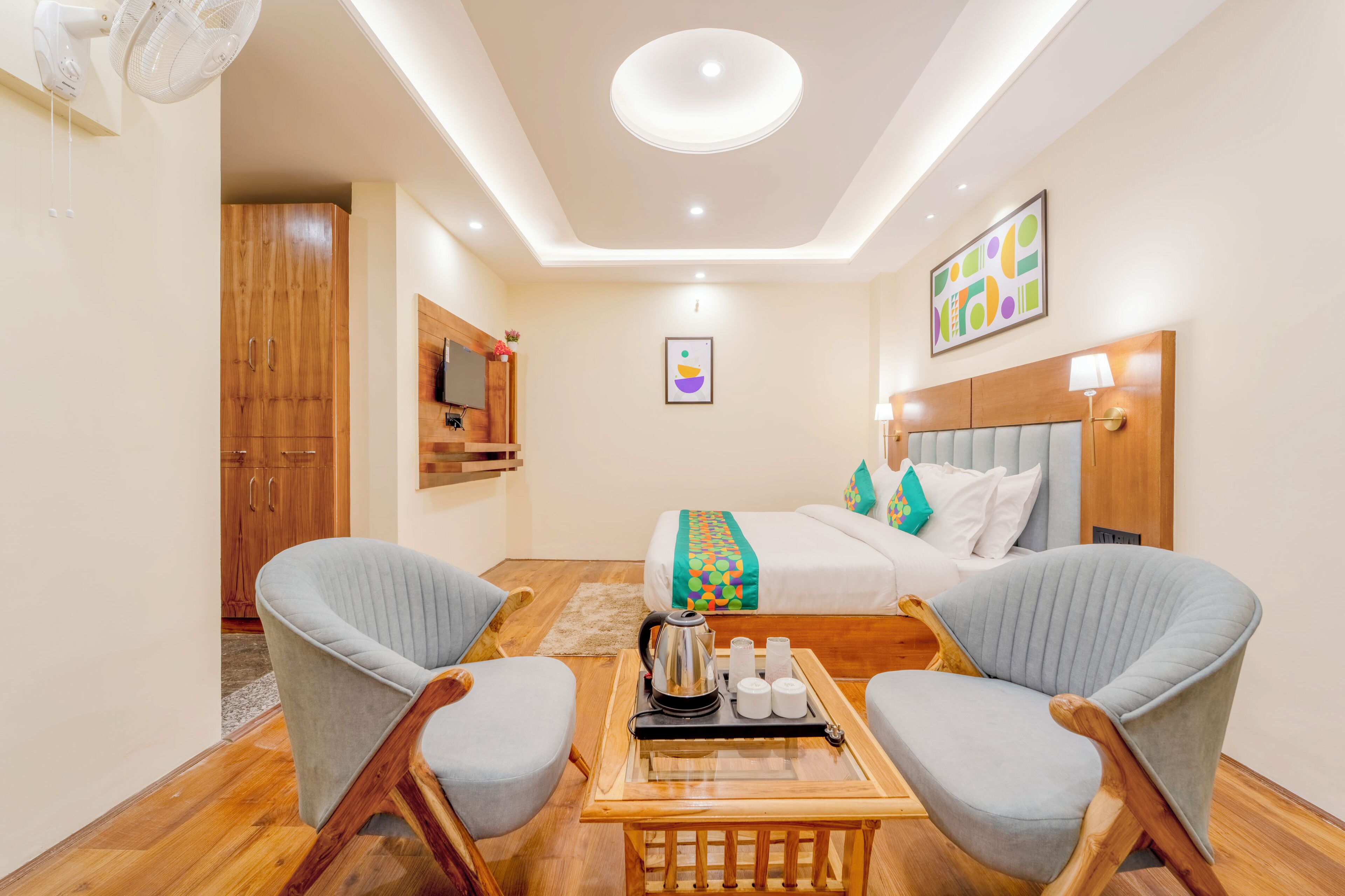 A hotel room with a bed, two armchairs, a wooden table with a tray, a ceiling fan, and a TV mounted on the wall.