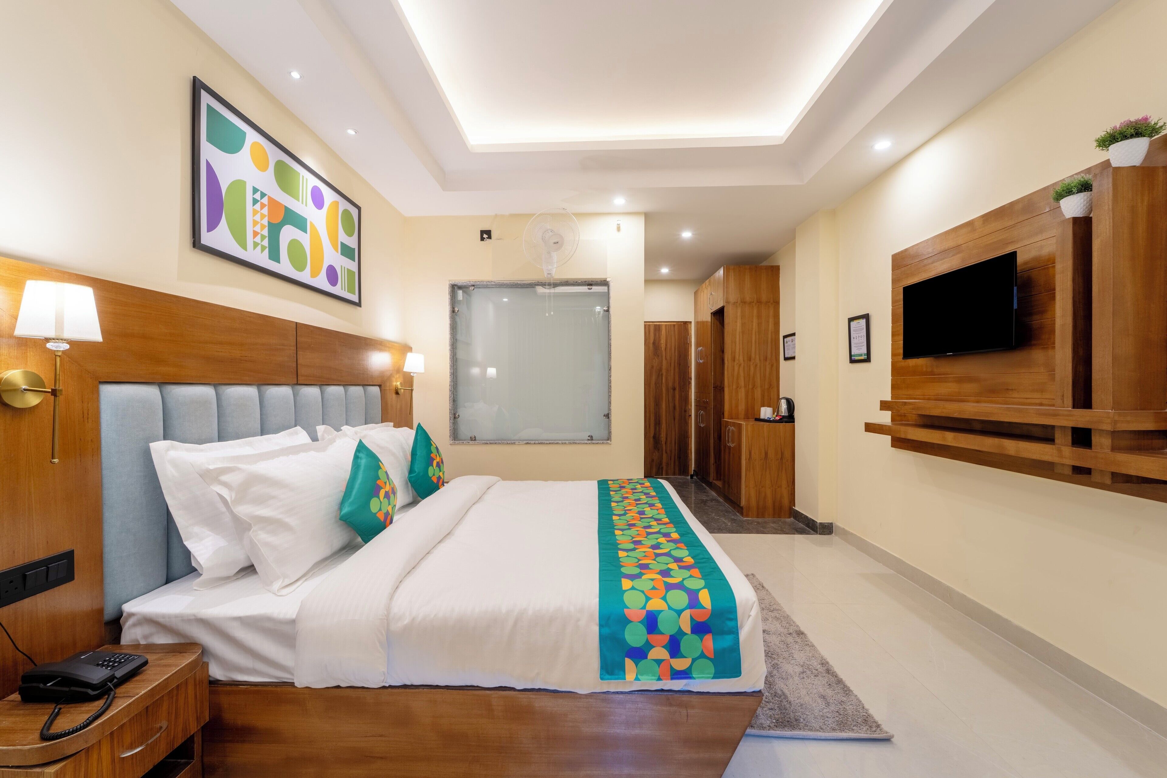 A hotel room with a bed, bedside table, lamp, TV, and a large window.