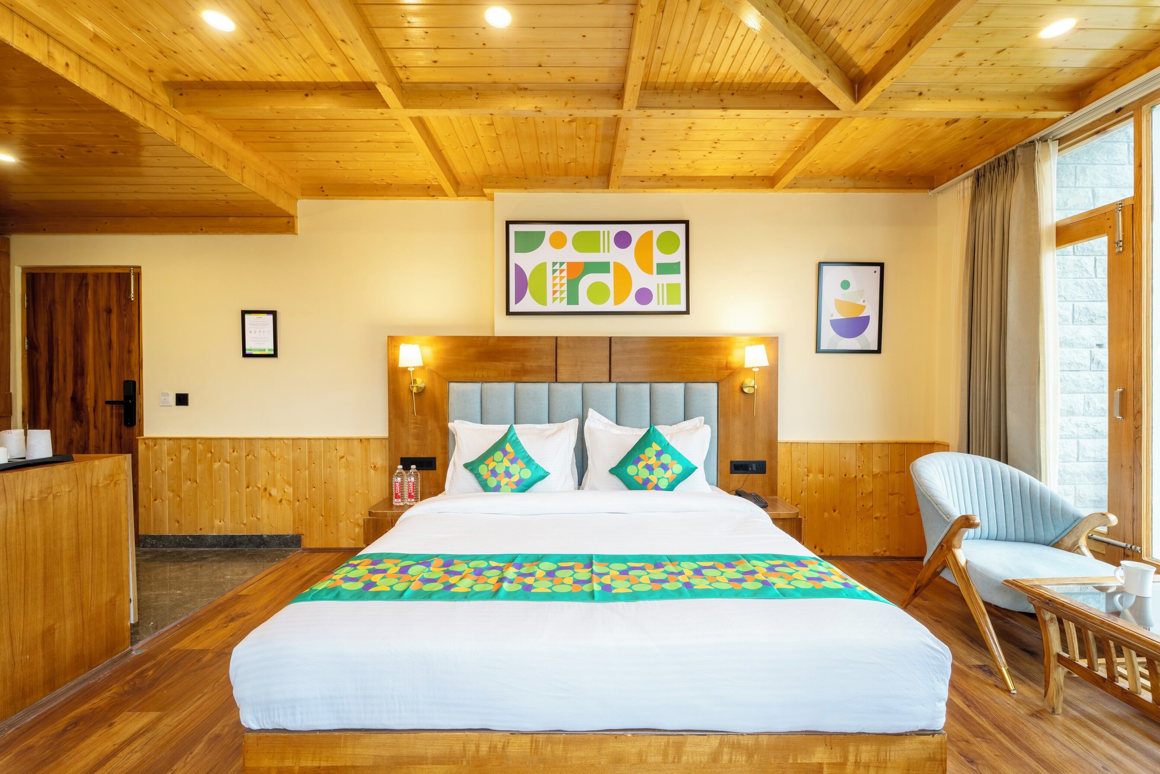 A hotel room with a wooden ceiling, a large bed with white bedding, two wall-mounted lamps, a wooden chair, and a window with curtains.