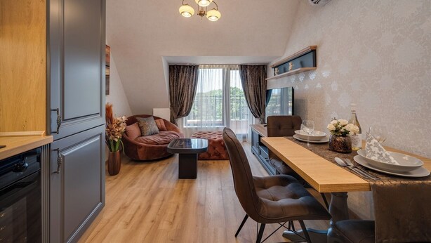 A modern kitchen with dark cabinets, a wooden countertop, and a dining area with a wooden table and chairs. - Prestige Central Apartment 10 (Varna)