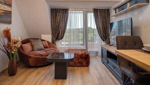A modern living room with a brown sofa, a tufted ottoman, a wooden coffee table, and a TV on a wooden stand. - Prestige Central Apartment 10 (Varna)