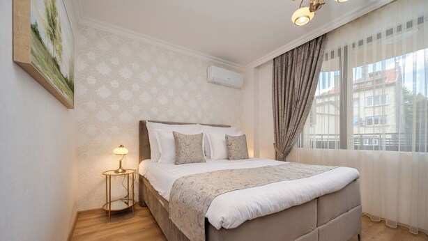 A neatly arranged bedroom with a large bed, a bedside table with a lamp, and a window with curtains.