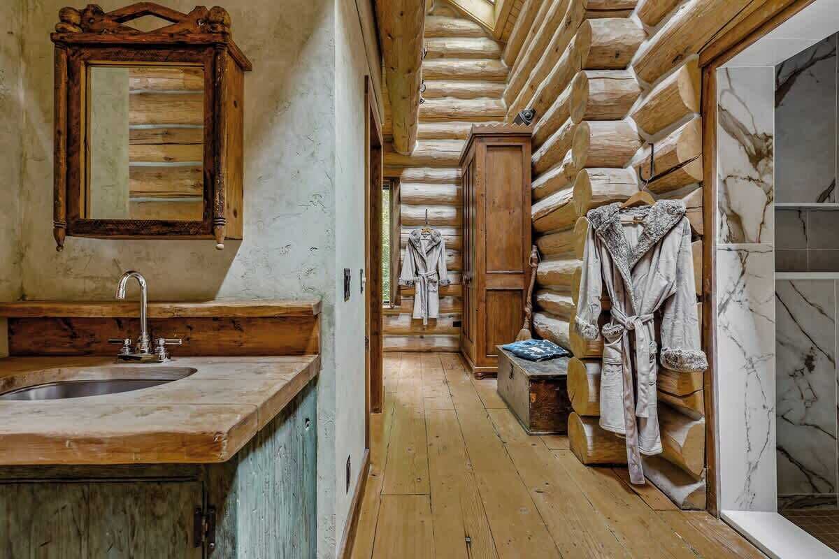 A log cabin interior with a wooden sink, a mirror, a robe rack, and a marble-tiled shower area.