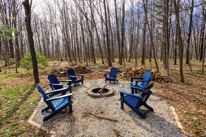 A forest setting with a fire pit and blue Adirondack chairs arranged around it.
