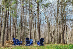 A forest with three blue Adirondack chairs and a stack of firewood.