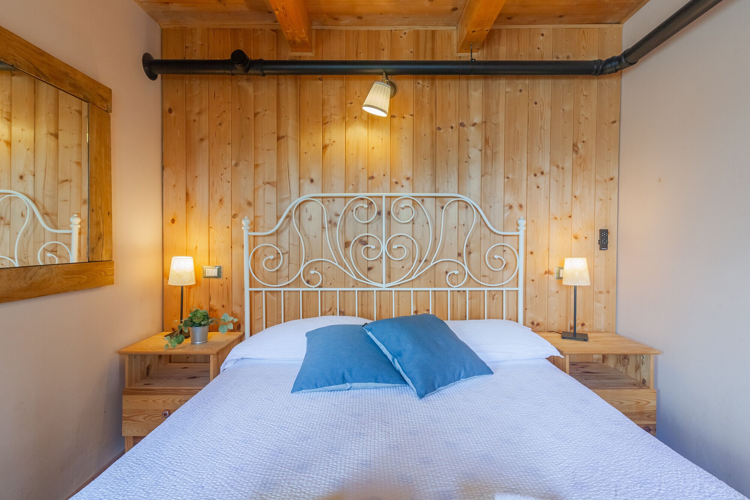 Ein Schlafzimmer mit einer holzverkleideten Wand, einem Metallbettgestell, zwei Nachttischen mit Lampen und einem großen Bett mit weißer Bettwäsche und blauen Kissen.