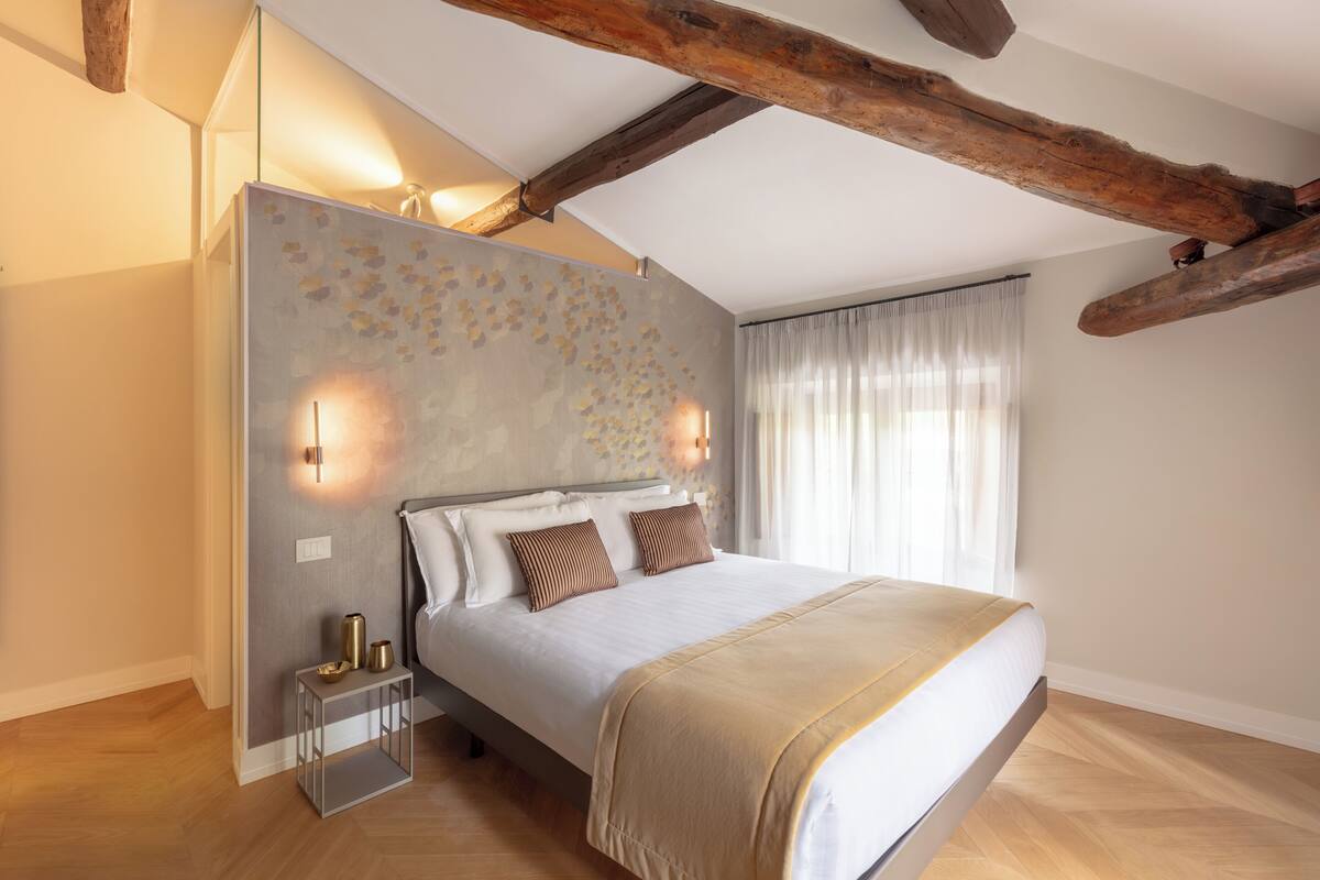 A bedroom with a bed, wooden beams, a wall with a textured pattern, and a window with curtains.
