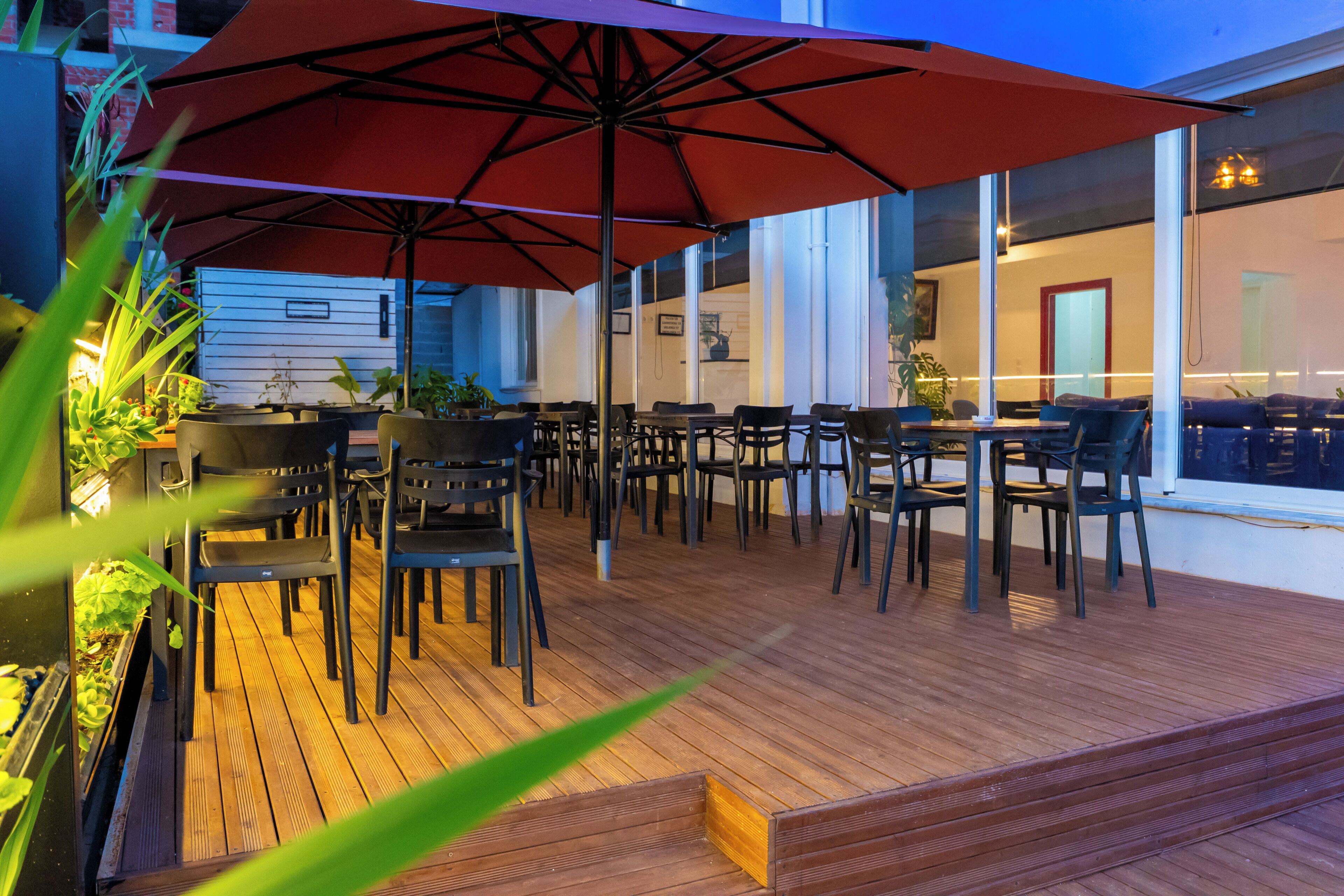 An outdoor patio with wooden flooring, multiple tables and chairs, and a large red umbrella providing shade.