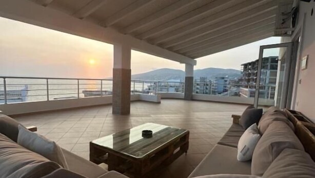 A spacious balcony with a sofa and a coffee table, overlooking a body of water and distant hills. - Alnes Hotel (Sarande)