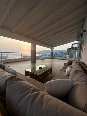 A spacious balcony with a sofa and a coffee table, overlooking a body of water and distant hills. - Alnes Hotel (Sarande)