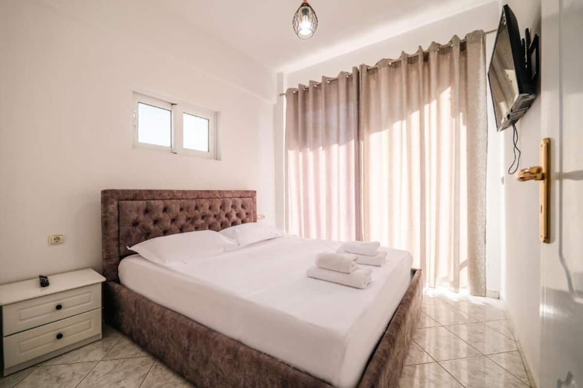 A bedroom with a large bed, a bedside table, a TV mounted on the wall, and a window with curtains.