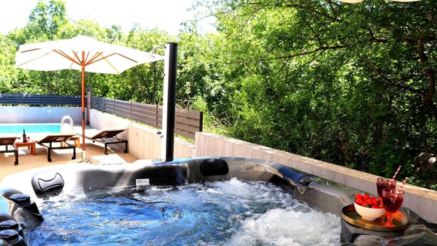 A hot tub with water jets, surrounded by a pool and lounge chairs, with a wooden deck and greenery in the background.